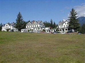 A large, multi-story building with a symmetrical facade, likely a hotel or resort, surrounded by a grassy field and tall trees. - Hotel Halsingland (Haines)
