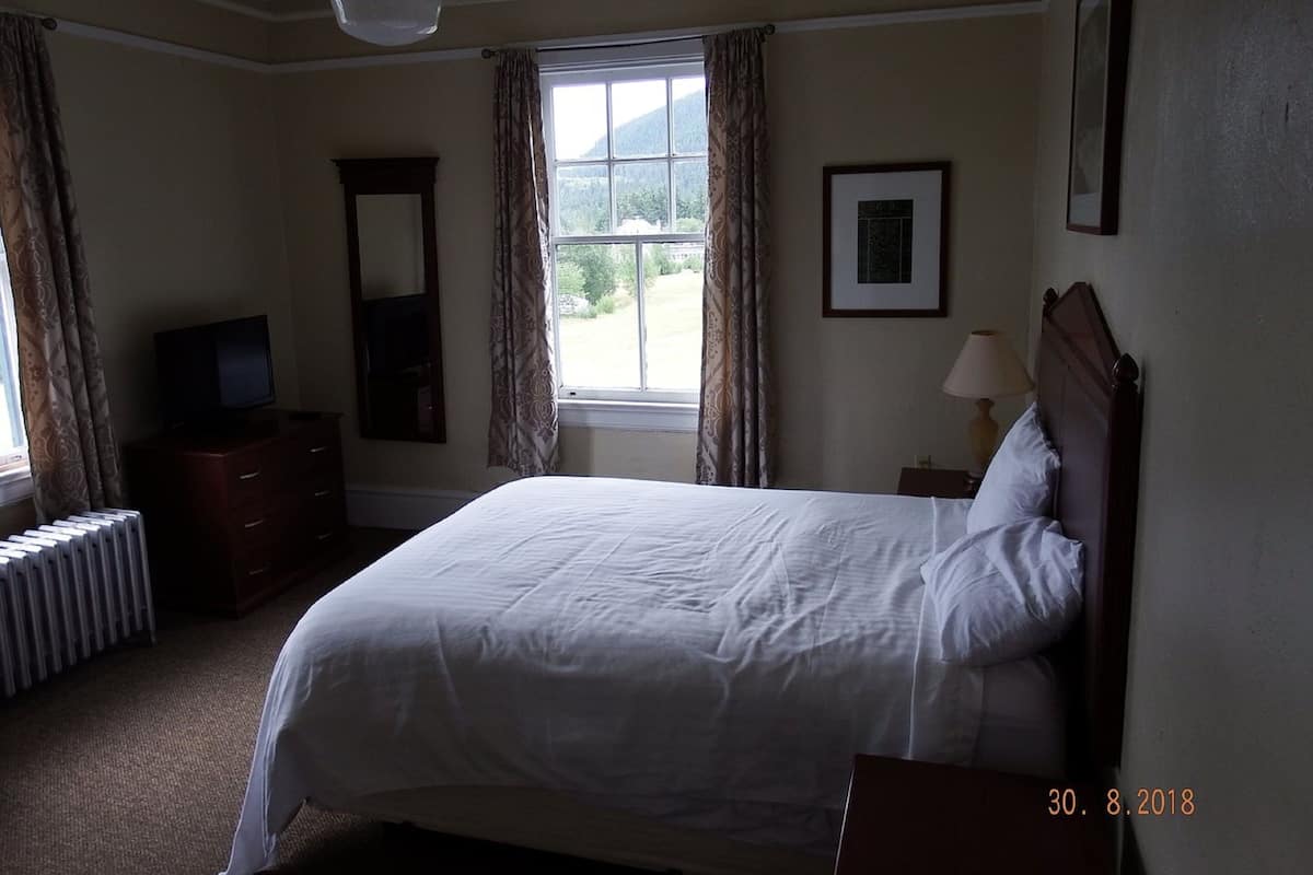 A hotel room with a bed, a dresser, a television, a window with curtains, a radiator, and a framed picture on the wall.