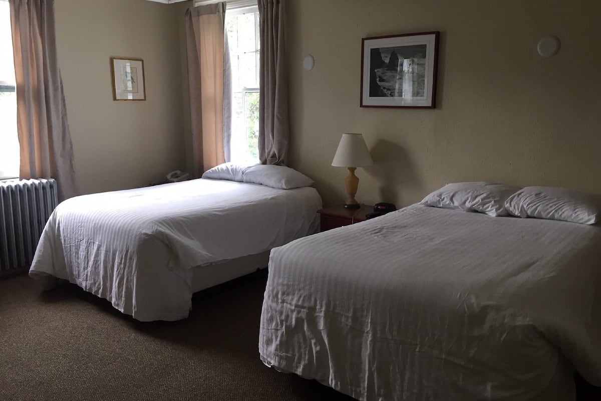 A hotel room with two beds, a window with curtains, a lamp, and a framed picture on the wall.