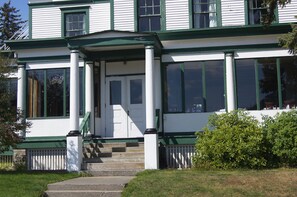 A two-story building with a green and white exterior, a front porch with steps, and a white picket fence. - Hotel Halsingland (Haines)
