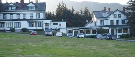 A large white building with multiple windows and a covered front porch, surrounded by a grassy field and parked cars.