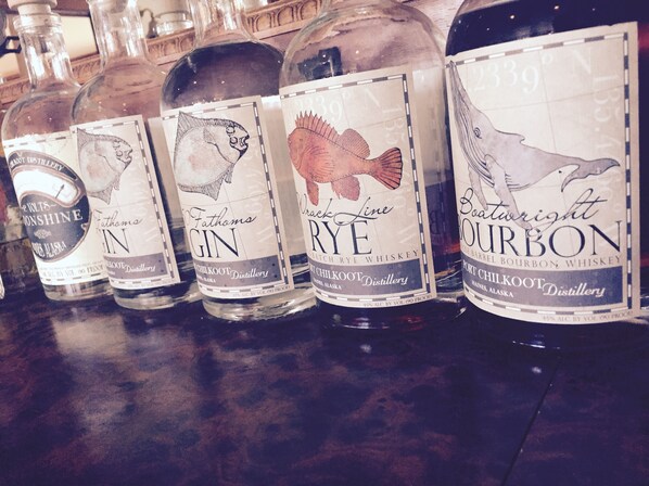 A row of whiskey bottles with labels, including 'Fathoms Gin', 'Fathoms Rye', and 'Port Chilkoot Bourbon'. - Hotel Halsingland (Haines)