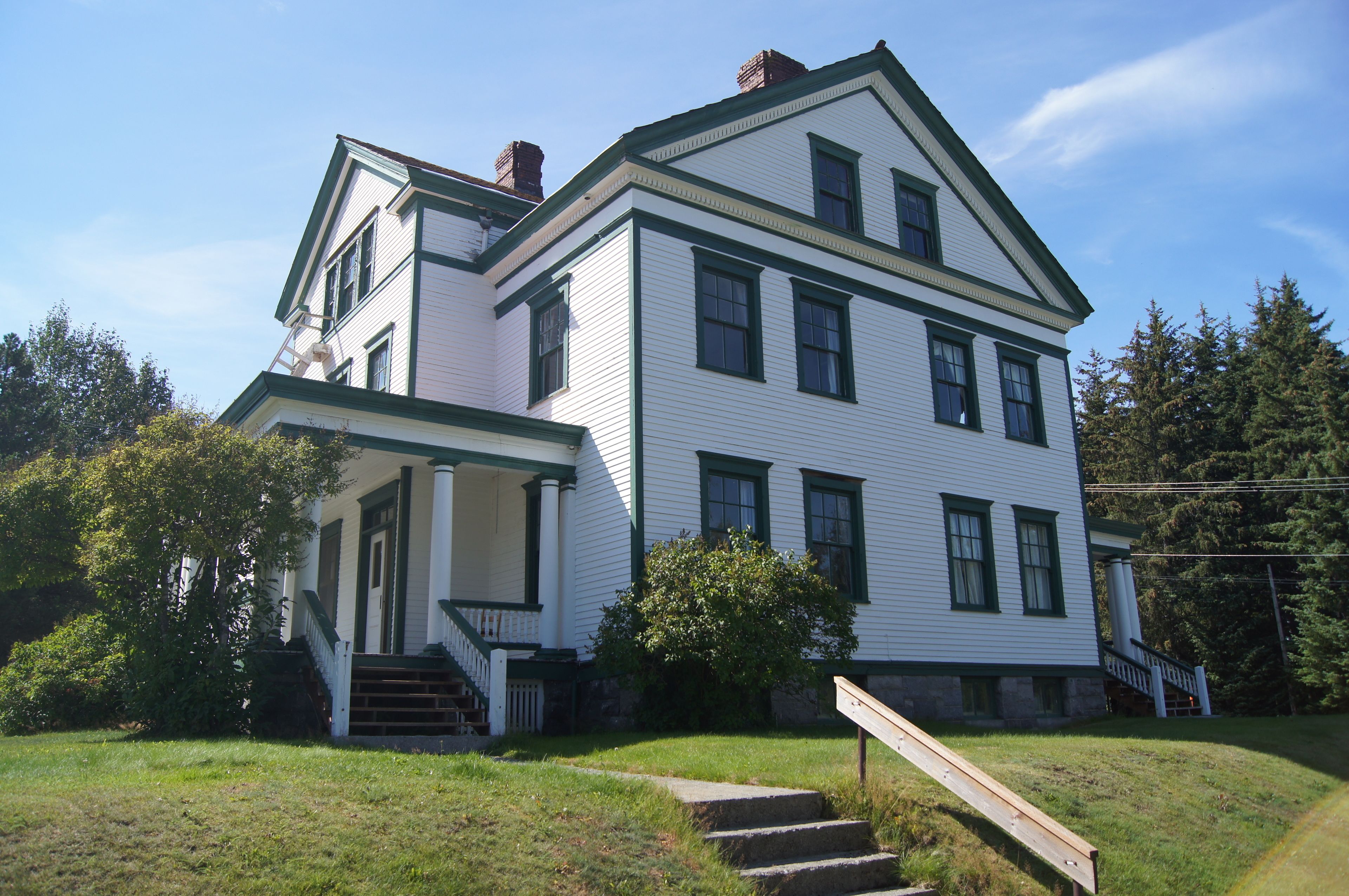 A two-story white house with green trim, a front porch, and a staircase leading to the entrance.