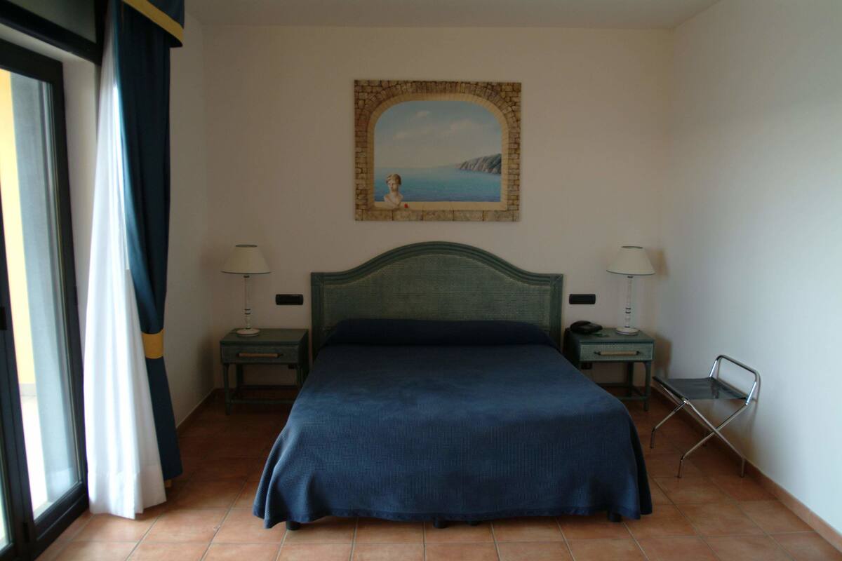 Une chambre à coucher avec un lit, deux tables de chevet, une commode, une chaise, un tableau au mur et une fenêtre avec des rideaux.