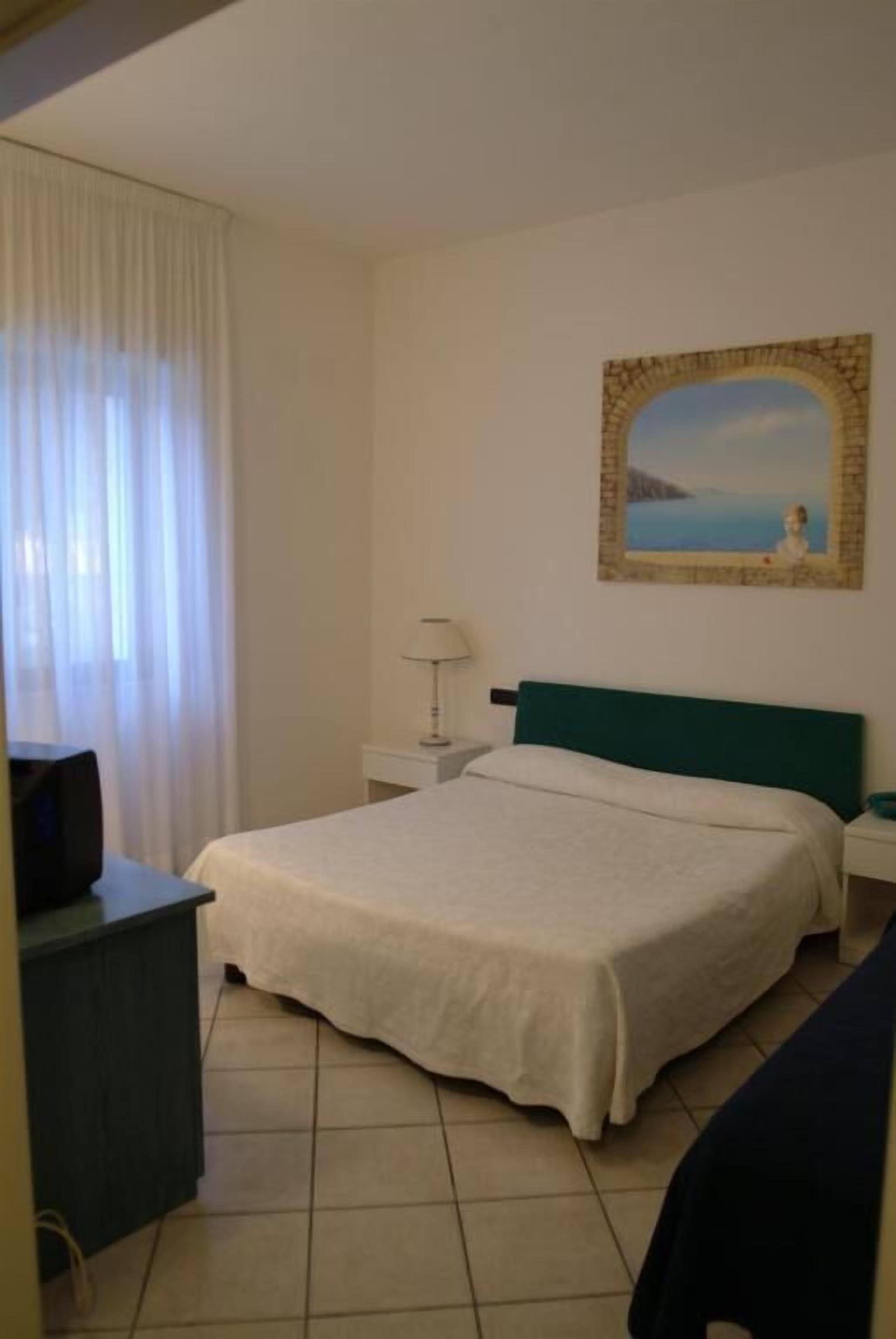 Hotelkamer met een bed, nachtkastje, lamp, schilderij aan de muur en een raam met gordijnen.