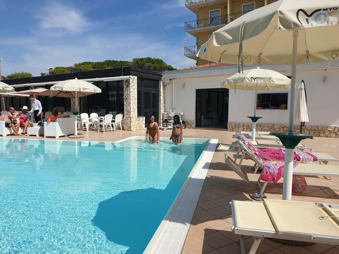 Un área de piscina con sillones de descanso y sombrillas, un edificio con balcones y cielo despejado.