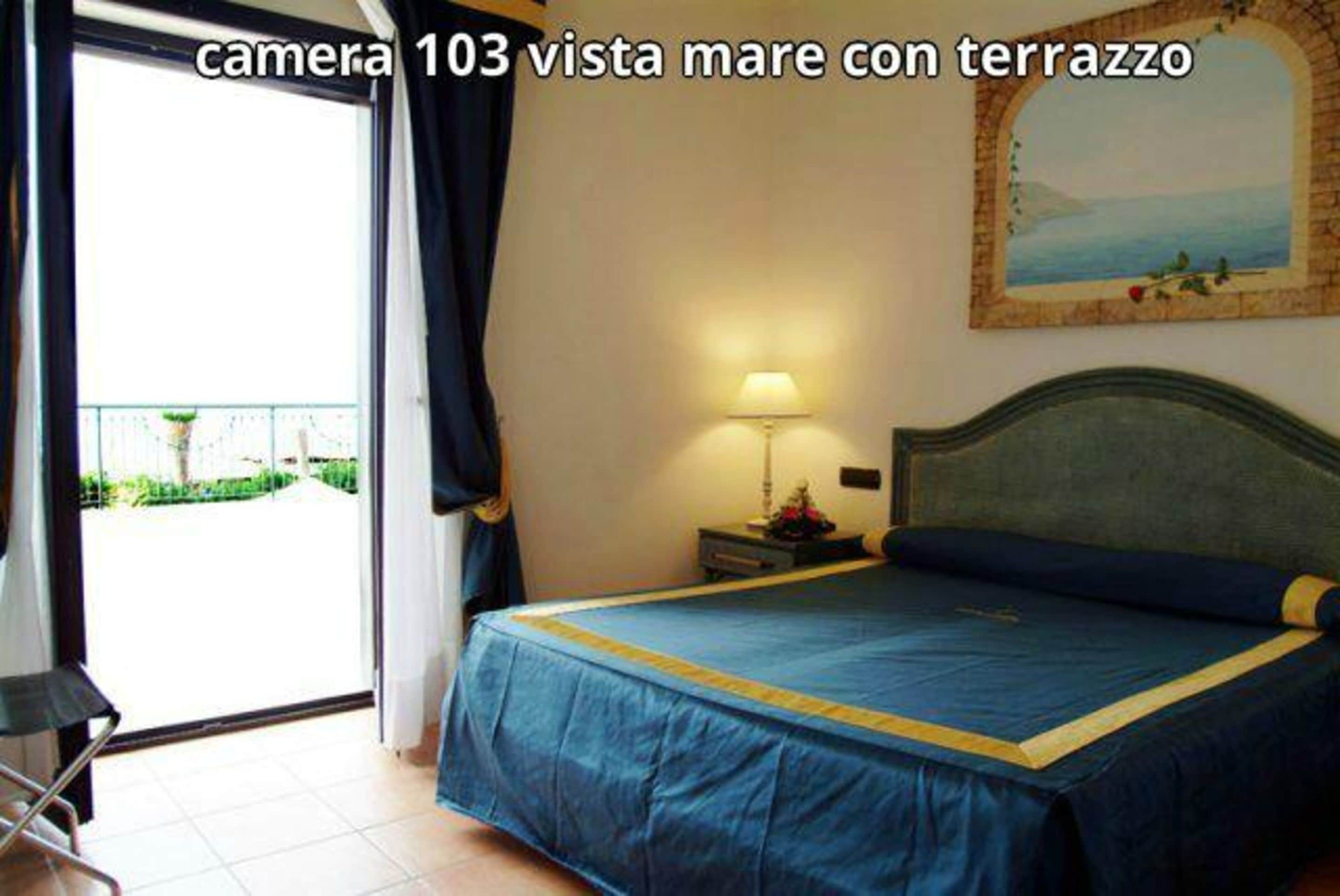 Habitación de hotel con cama, mesita de noche, lámpara, un cuadro y vista al mar a través de una ventana.