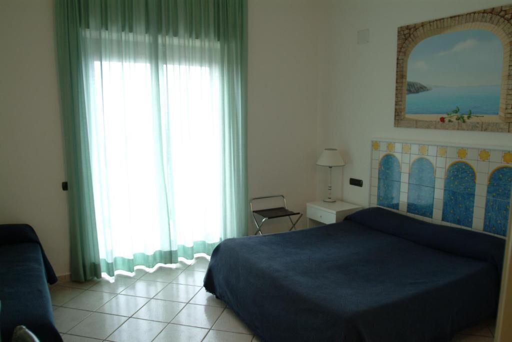 A hotel room with two beds, a window with curtains, a small table with a lamp, and a decorative wall tile.