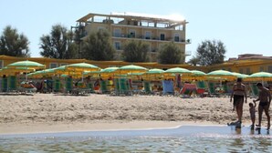 Une plage bordée de rangées de parasols verts et jaunes, une étendue de sable et, au loin, un immeuble à plusieurs étages.