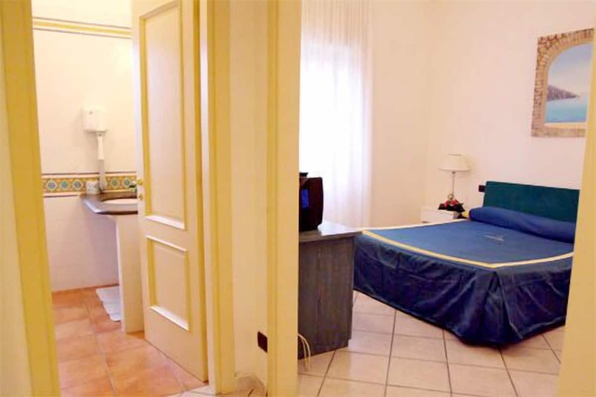 Une chambre d’hôtel avec un lit, un bureau avec un ordinateur, une fenêtre avec des rideaux et une salle de bain visible à travers une porte ouverte.