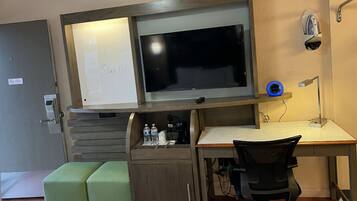 A compact hotel room with a desk, a green cushioned bench, a flat-screen TV, a small shelf with toiletries, and a security camera.