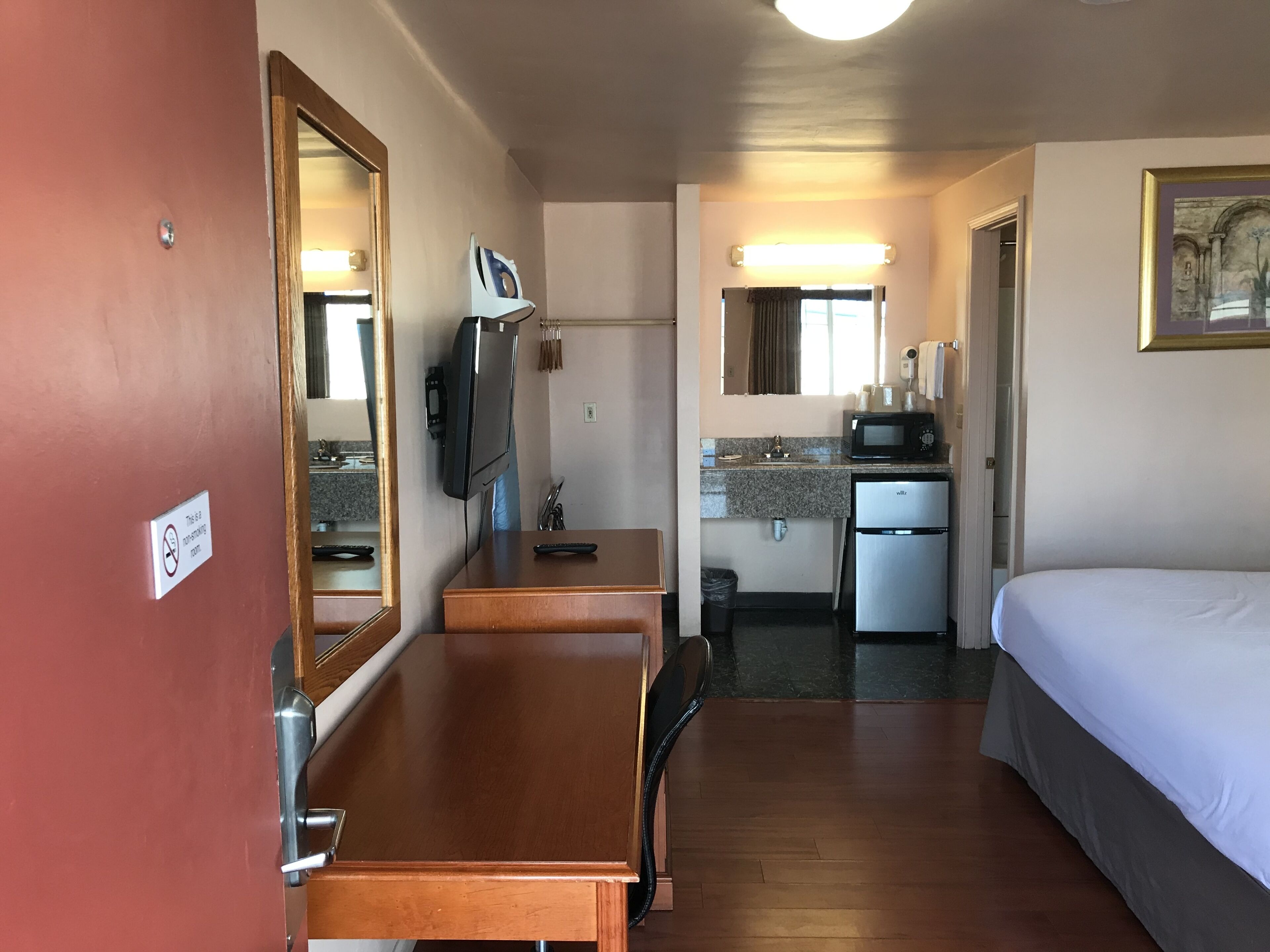 A compact hotel room with a wooden desk, a bed, a kitchenette with a microwave, and a large mirror.