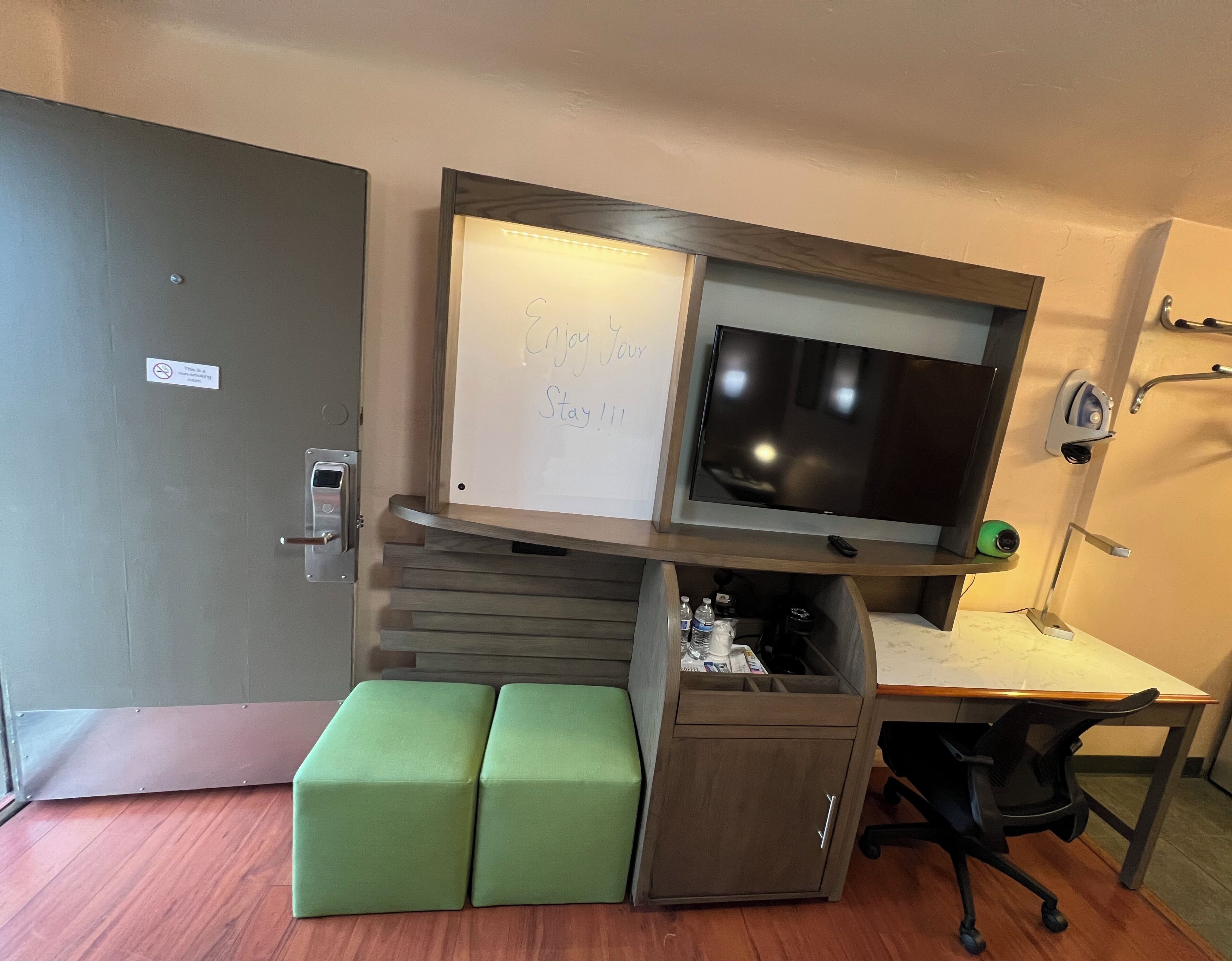 Habitació d'hotel petita amb escriptori, televisió de pantalla plana, cuineta compacta i un pouf verd.