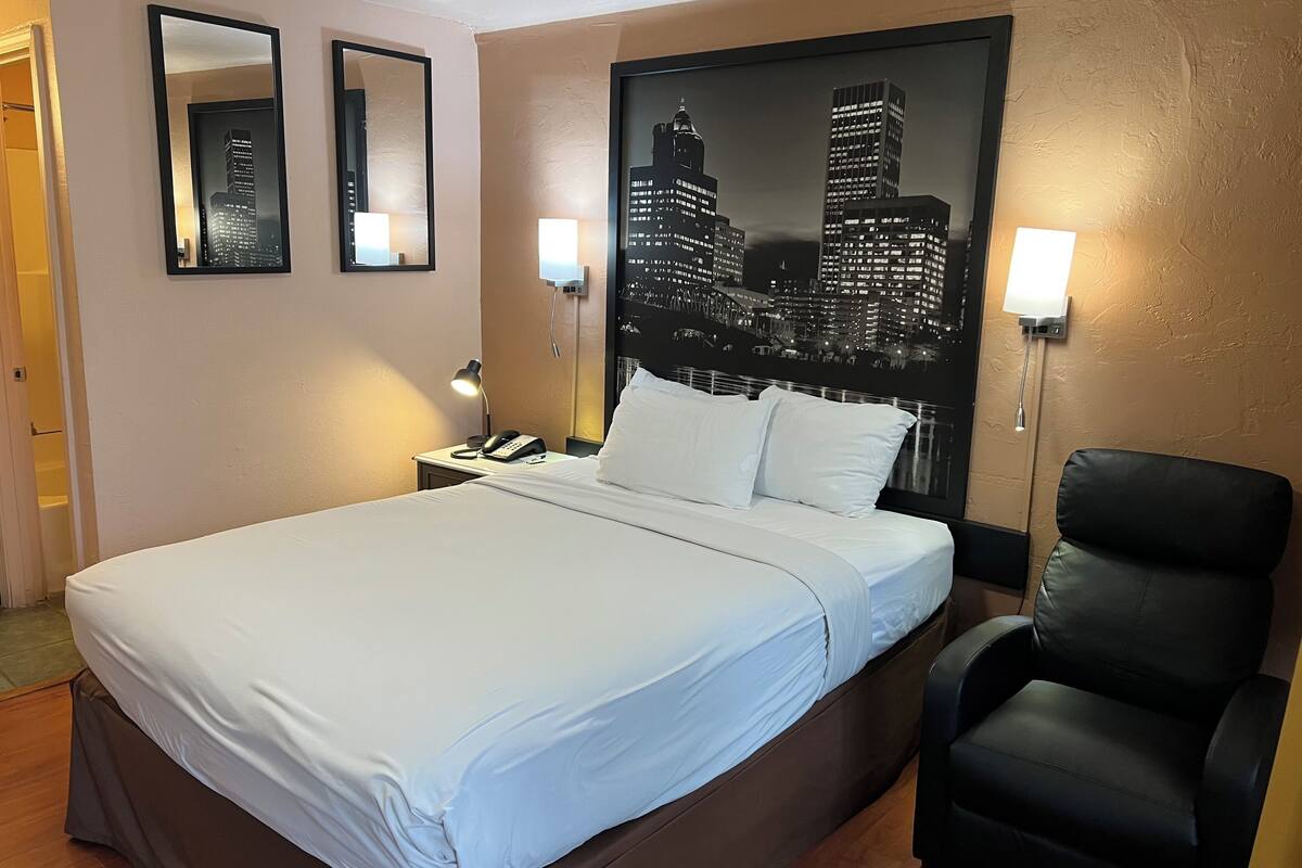 Ein Hotelzimmer mit einem großen Bett, zwei Nachttischlampen, einem schwarzen Ledersessel und einem Stadtbild an der Wand.