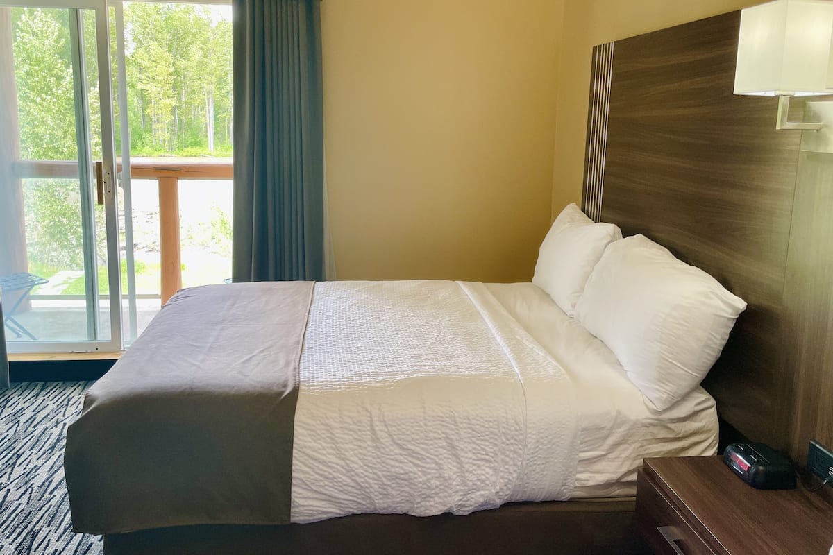 Quarto de hotel com uma cama grande, cabeceira de madeira, mesa de cabeceira e vista para uma varanda com cortinas.