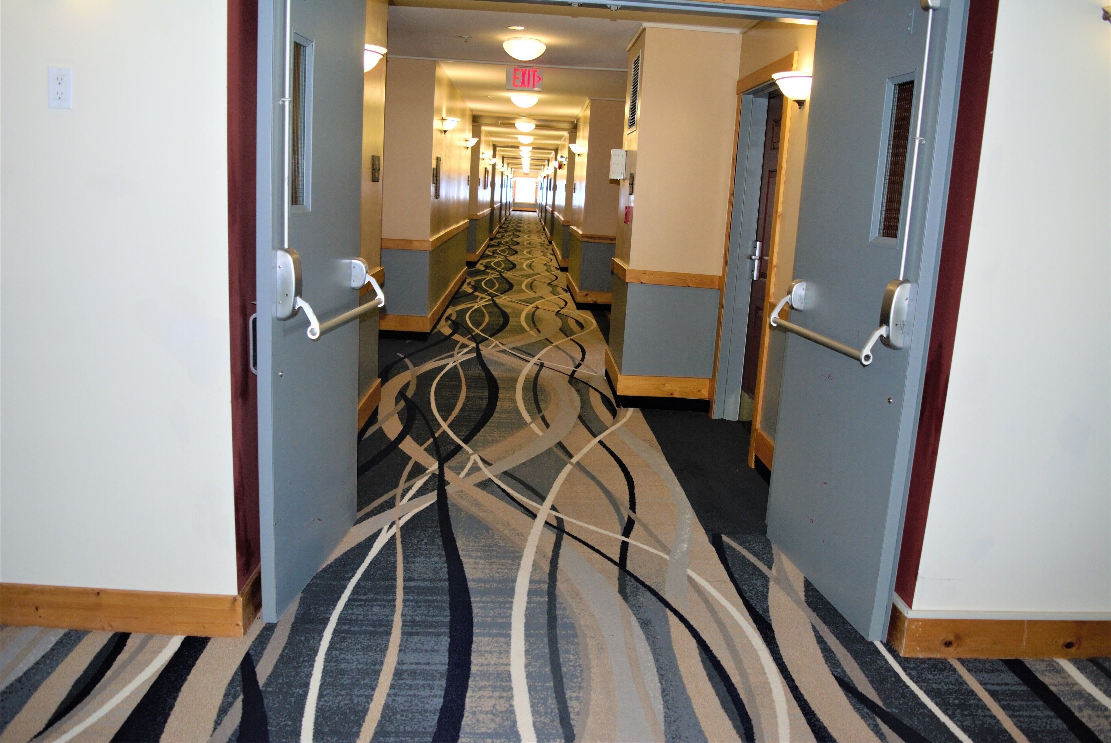 A hotel hallway with a patterned carpet, double doors, and a long corridor leading to a bright exit area.