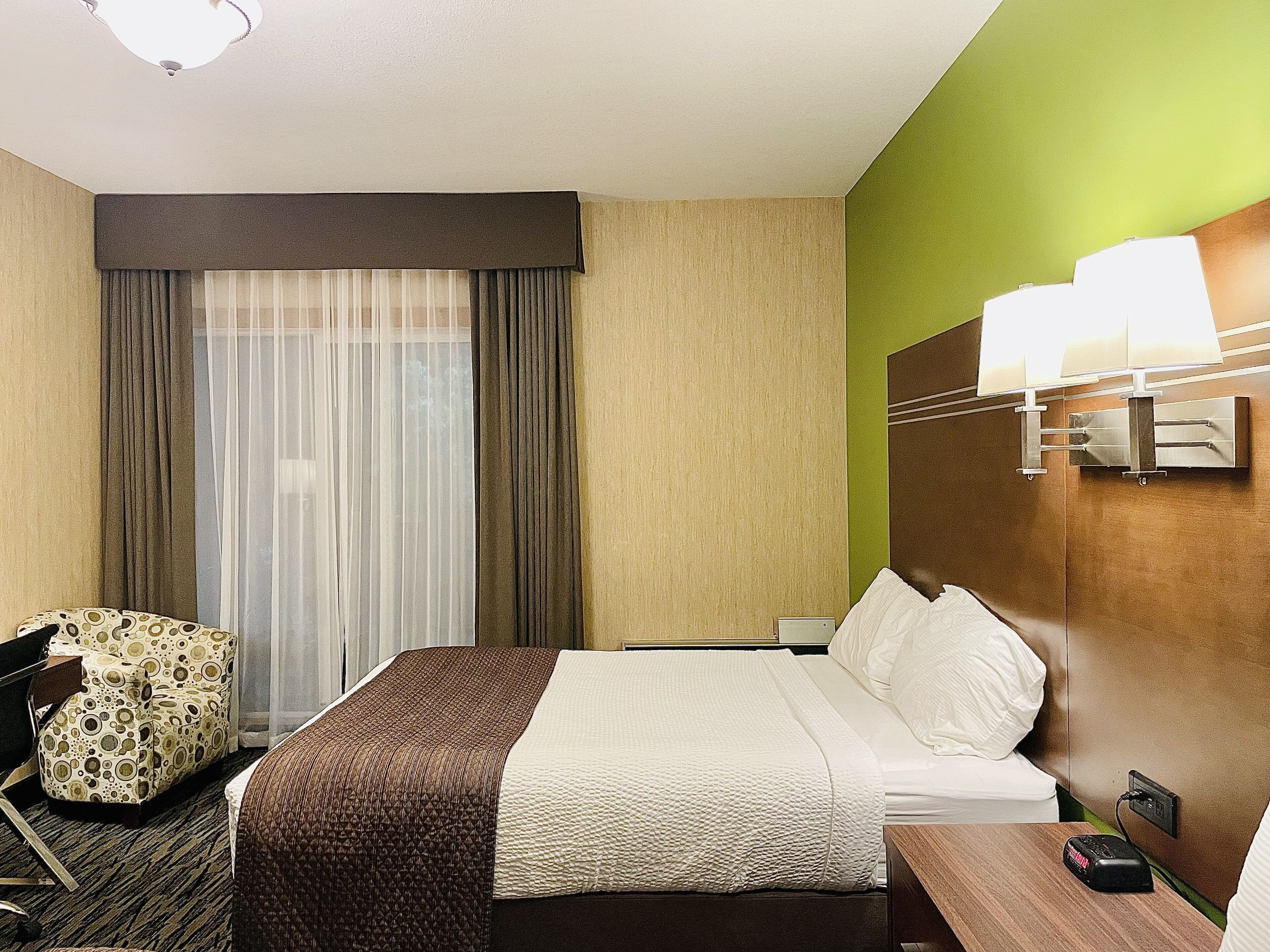 A hotel room with a bed, a chair, a desk, a lamp, and a window with curtains.