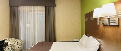 A hotel room with a bed, a chair, a desk, a lamp, and a window with curtains.