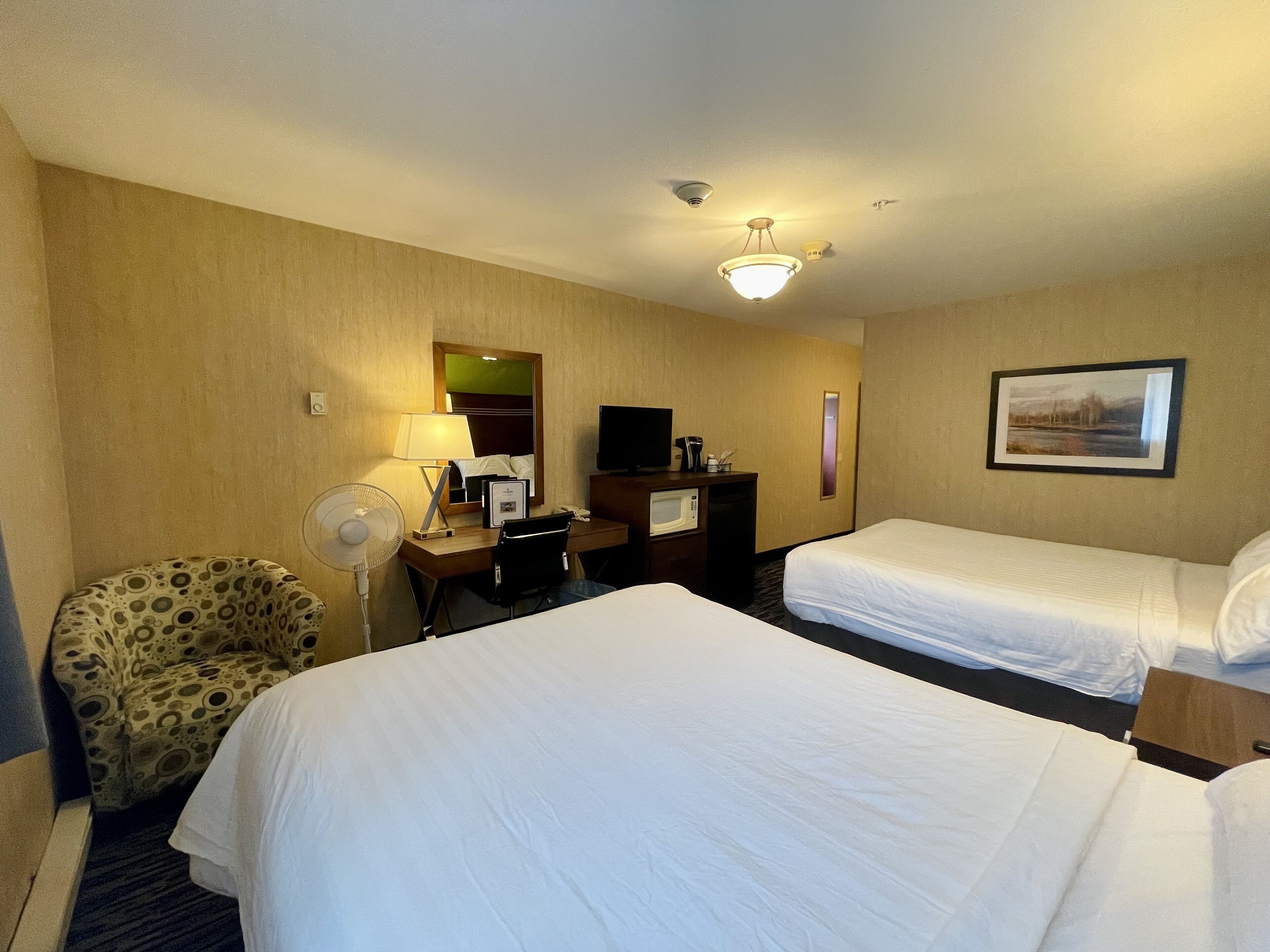 A hotel room with two beds, a desk with a computer, a chair, a fan, and a picture on the wall.