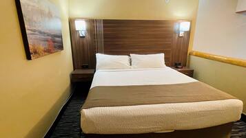 Une chambre d’hôtel avec un grand lit, deux tables de chevet, une petite table, une lampe fixée au mur et un tableau accroché au mur.