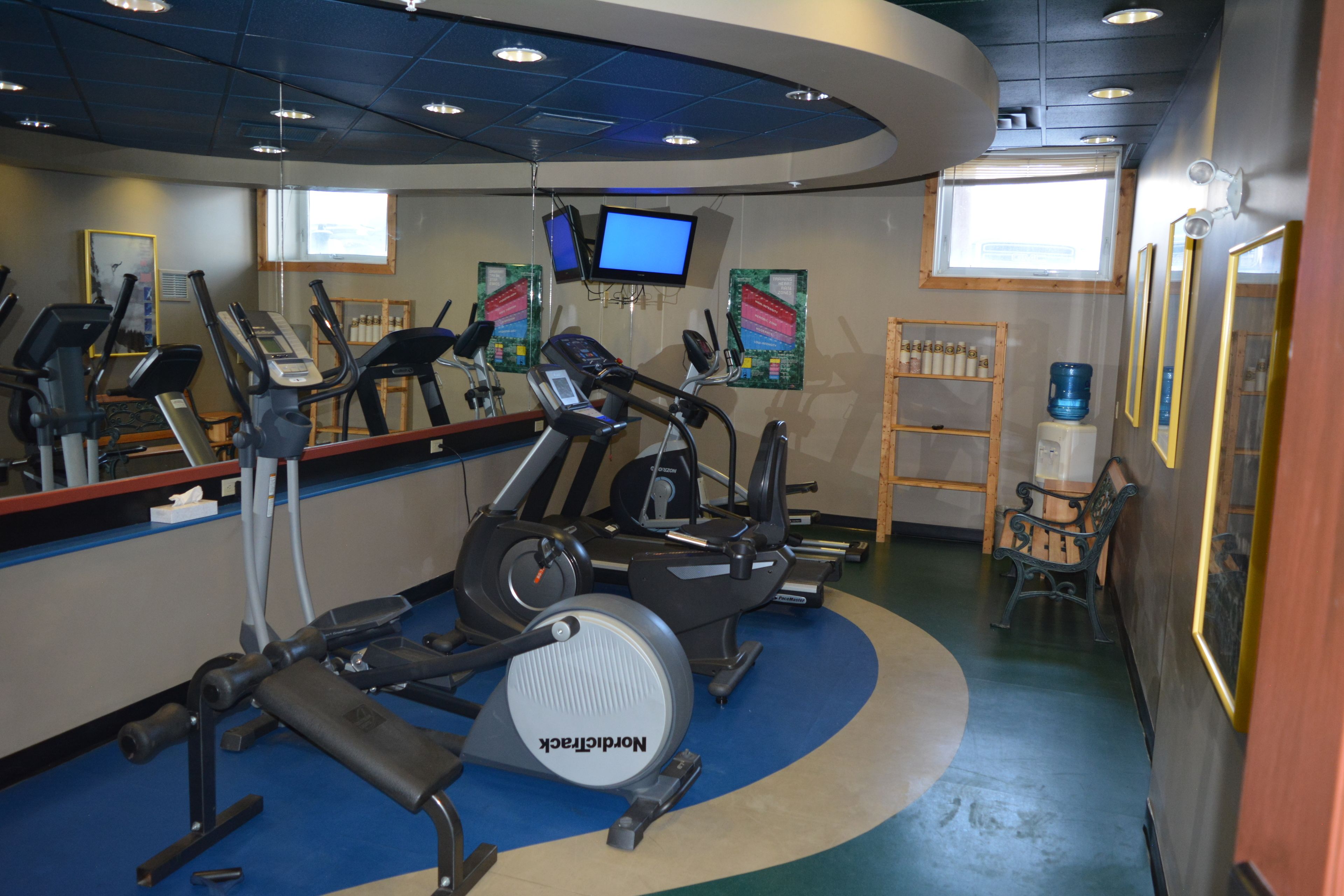 A gym with treadmills, stationary bikes, and a rowing machine. There are two monitors on the wall, a water cooler, and a bench.