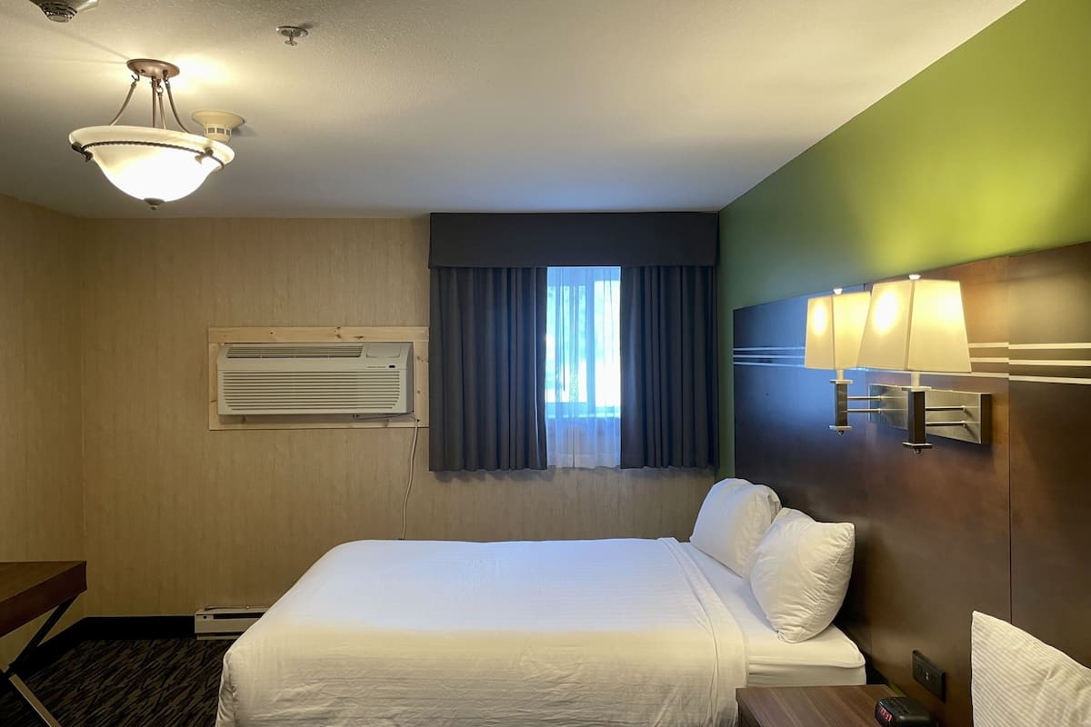 Quarto de hotel com cama, janela com cortinas, ar condicionado, luminária na parede e mesa de cabeceira com controle remoto.