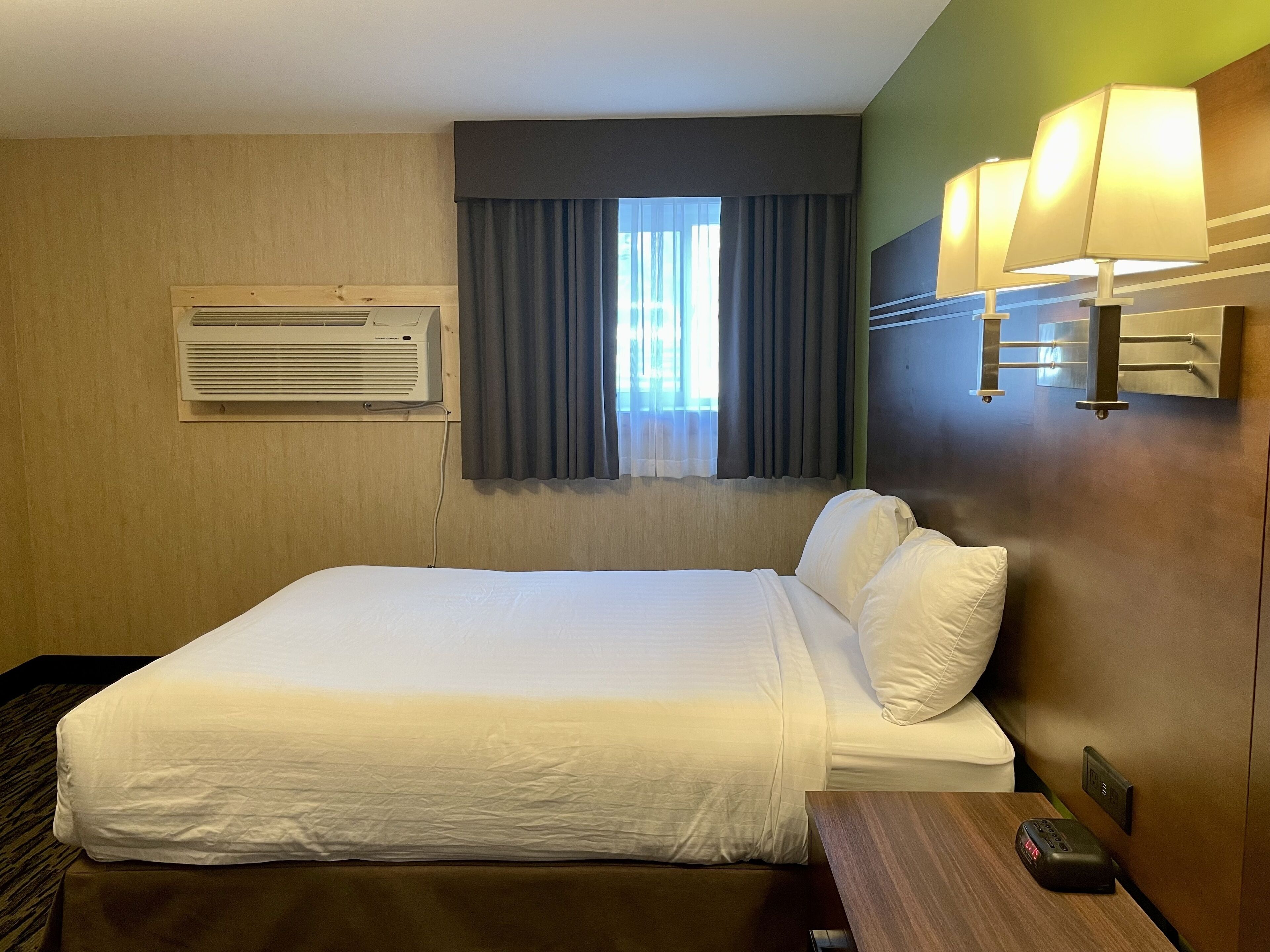 Una habitación de hotel con cama, ventana con cortinas, unidad de aire acondicionado, mesita de noche con lámpara y panel de control en la pared.