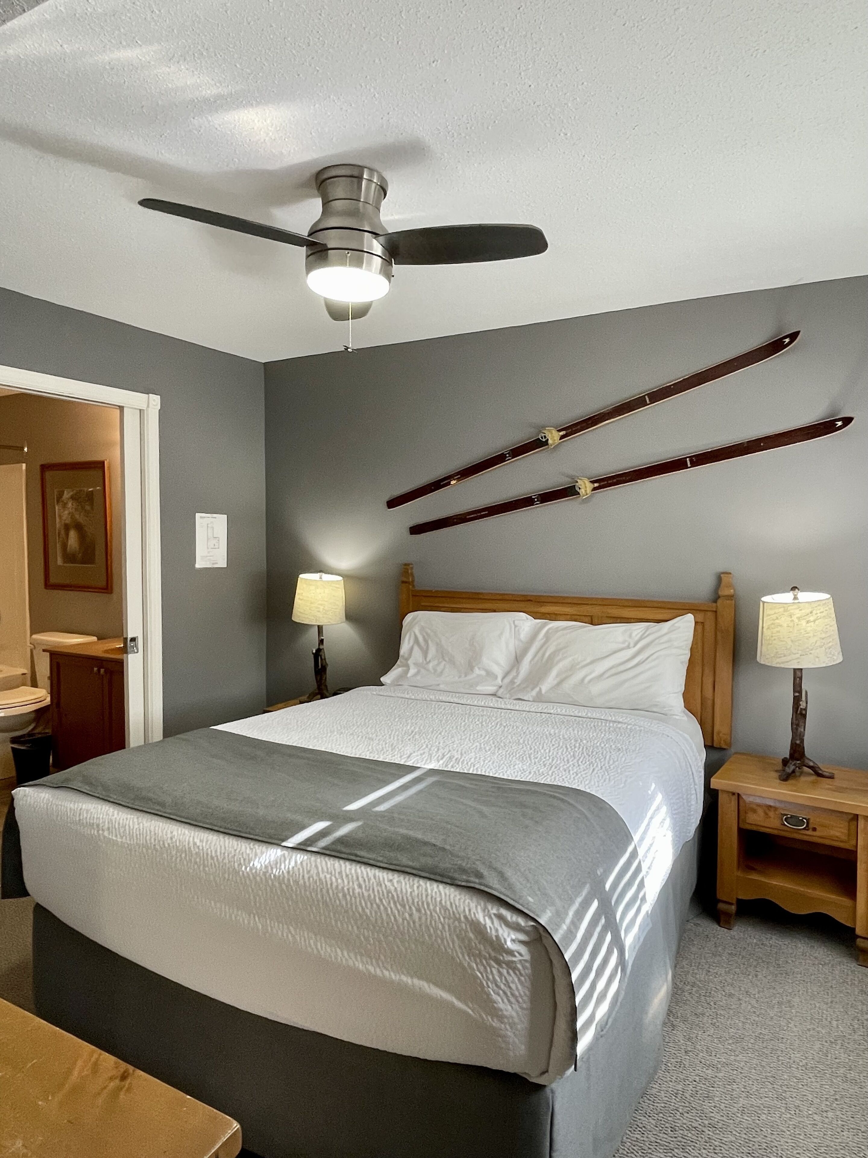A bedroom with a bed, two bedside tables with lamps, a ceiling fan, and a door leading to a bathroom.
