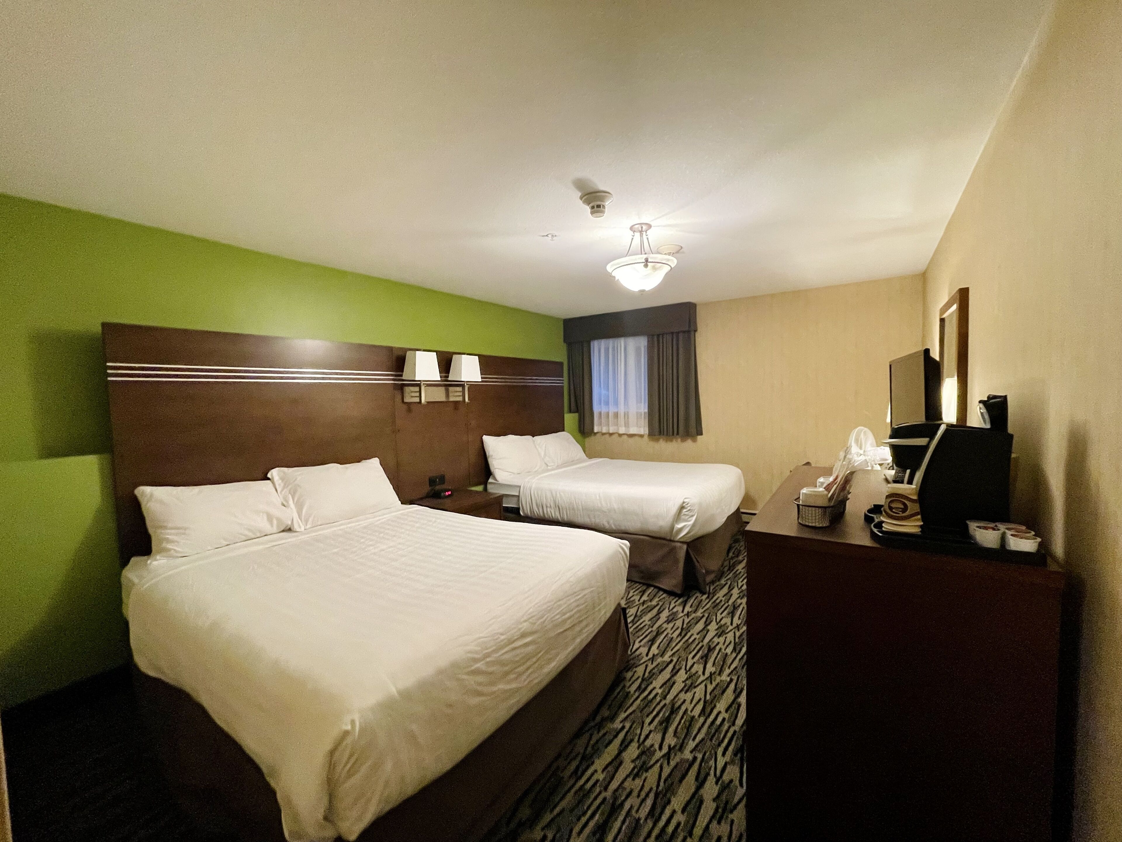A hotel room with two beds, a desk with a chair, a ceiling fan, and a window with curtains.
