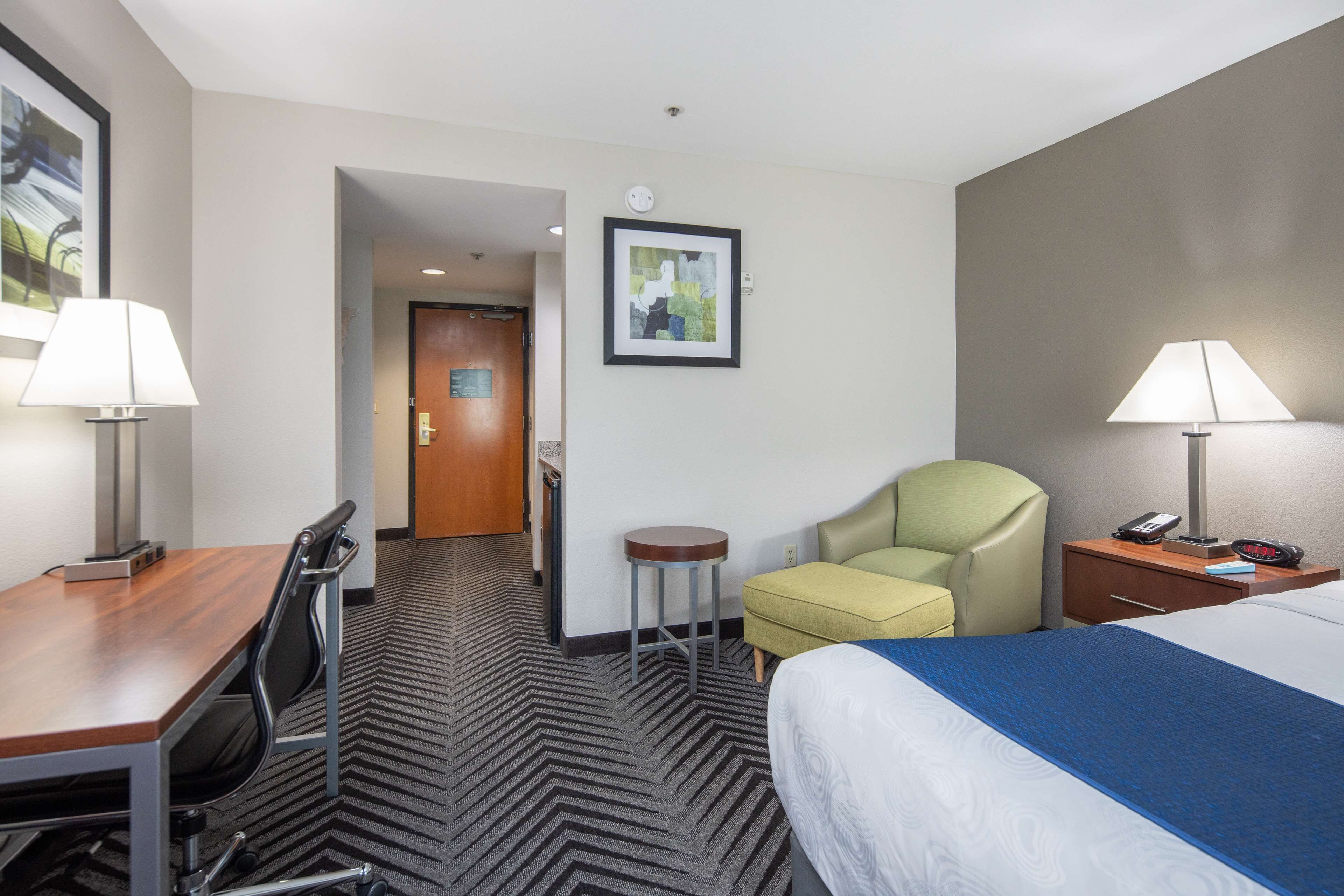 Quarto de hotel com cama, escrivaninha, cadeira, luminária e uma pequena mesa.