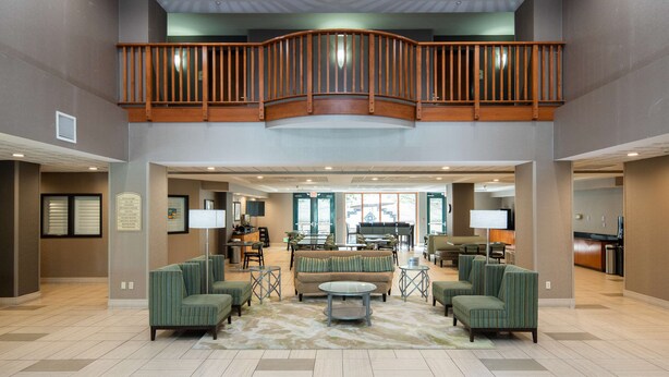Un hall d’entrée spacieux avec un plafond haut, un balcon en bois aux lignes courbes et un espace de détente central agrémenté de canapés et d’une table basse.