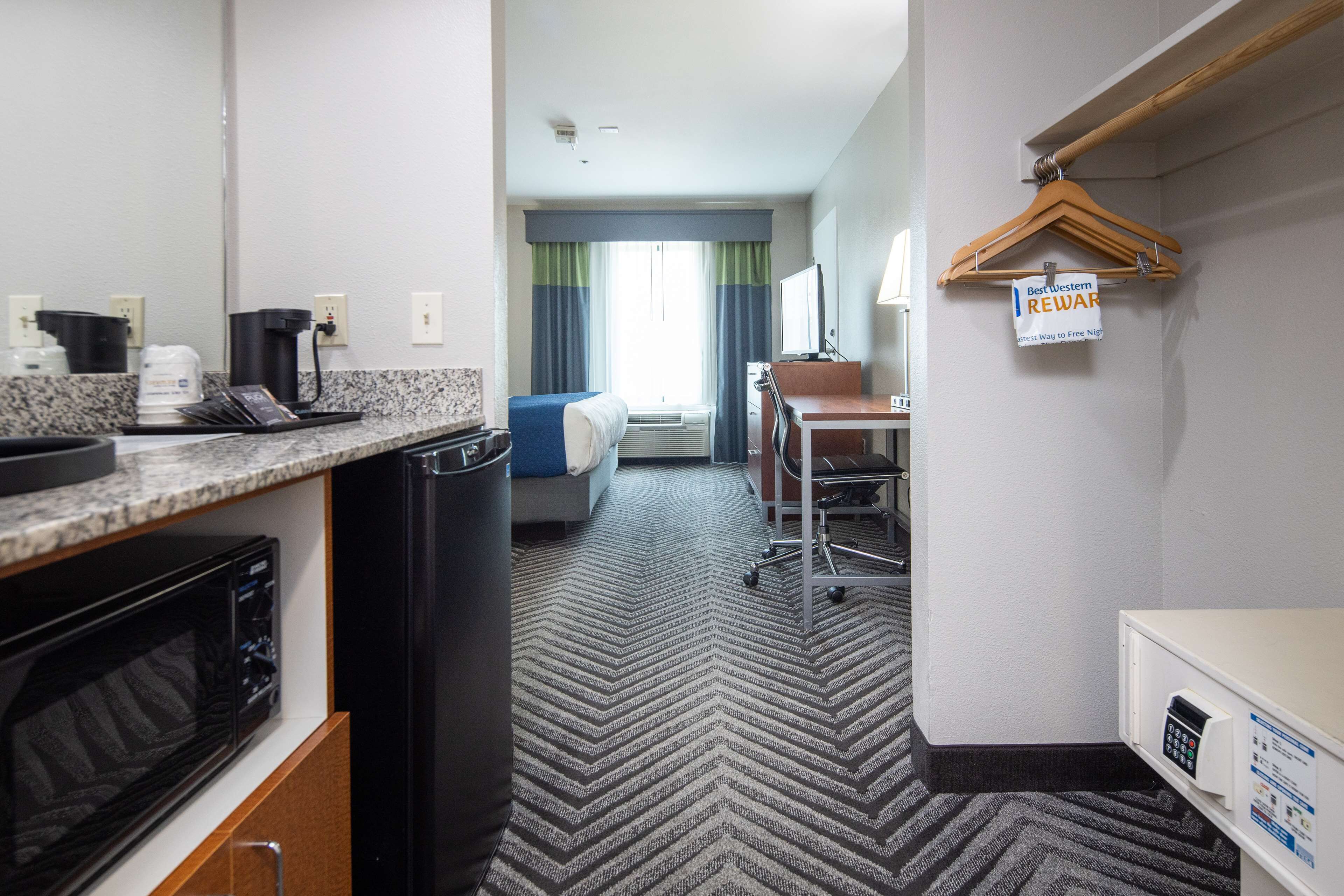 A hotel room with a kitchenette, a bed with a blue headboard, a desk with a chair, and a sign indicating a reward program.