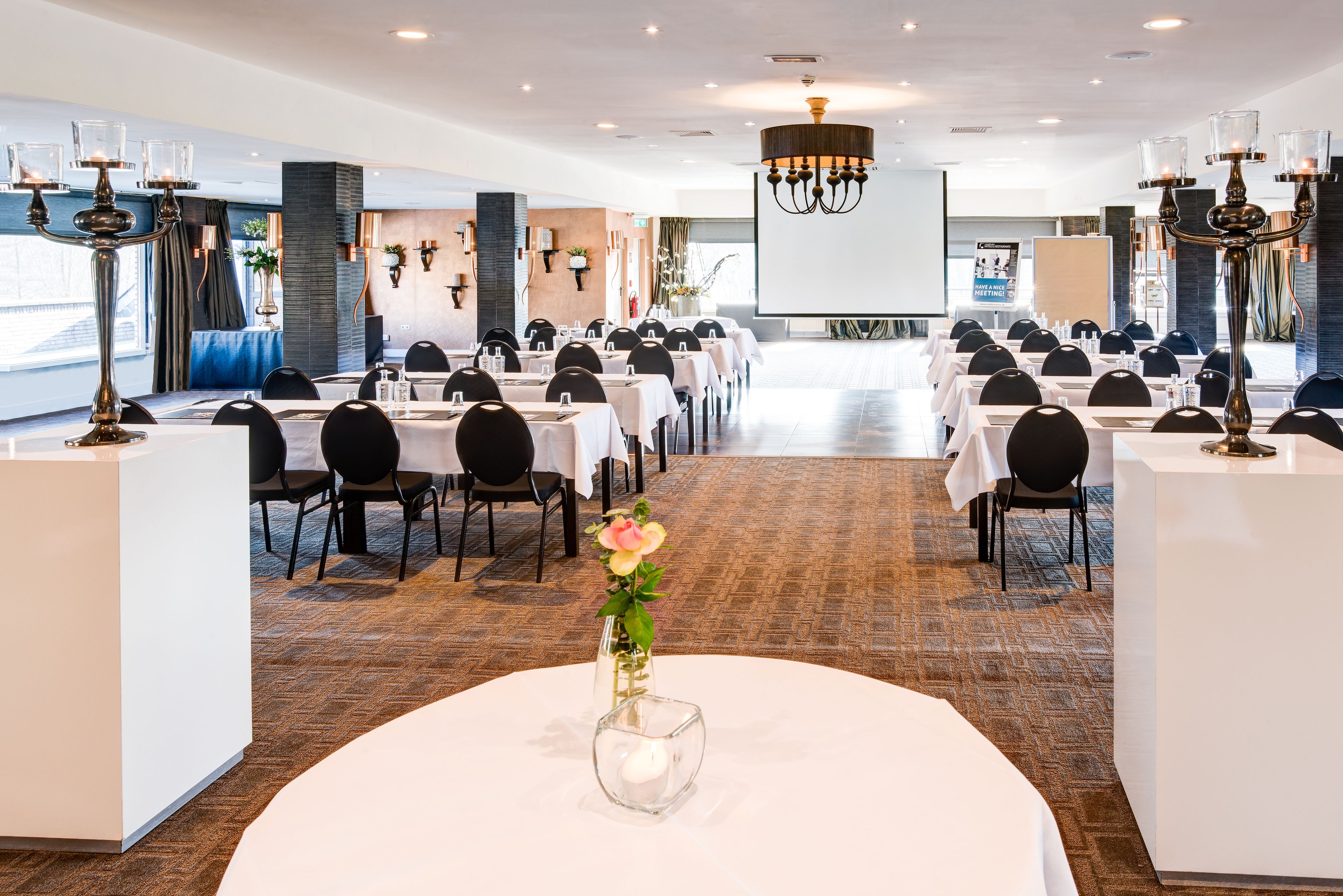 Un salón de conferencias con varias mesas y sillas, una pantalla grande y candelabros decorativos.