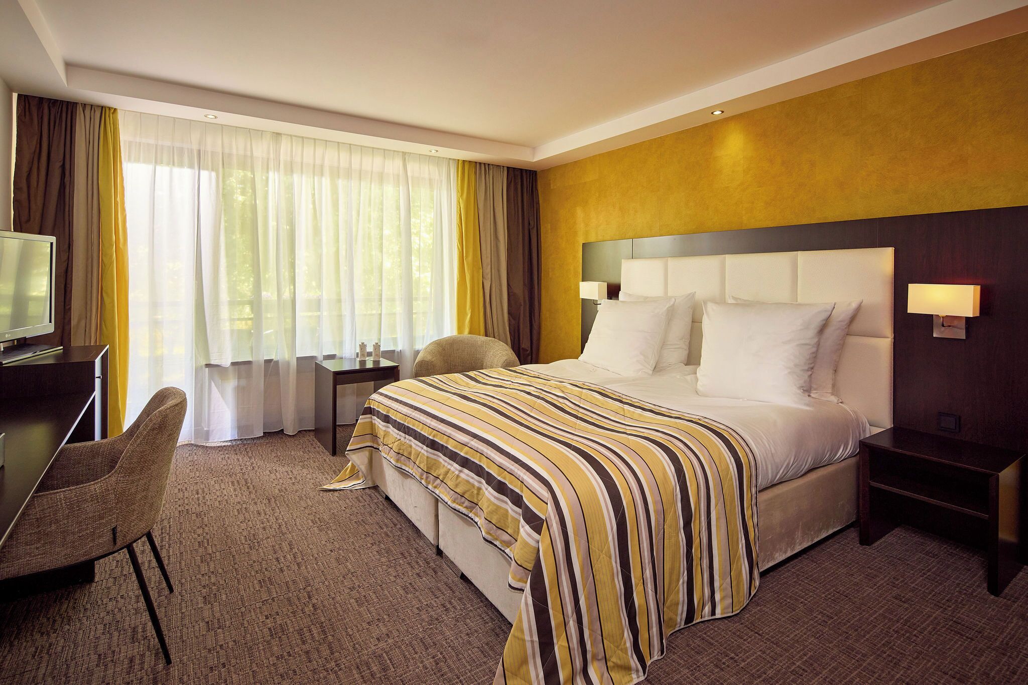 Une chambre d’hôtel avec un lit, un bureau, une chaise, une télévision et une fenêtre avec des rideaux.
