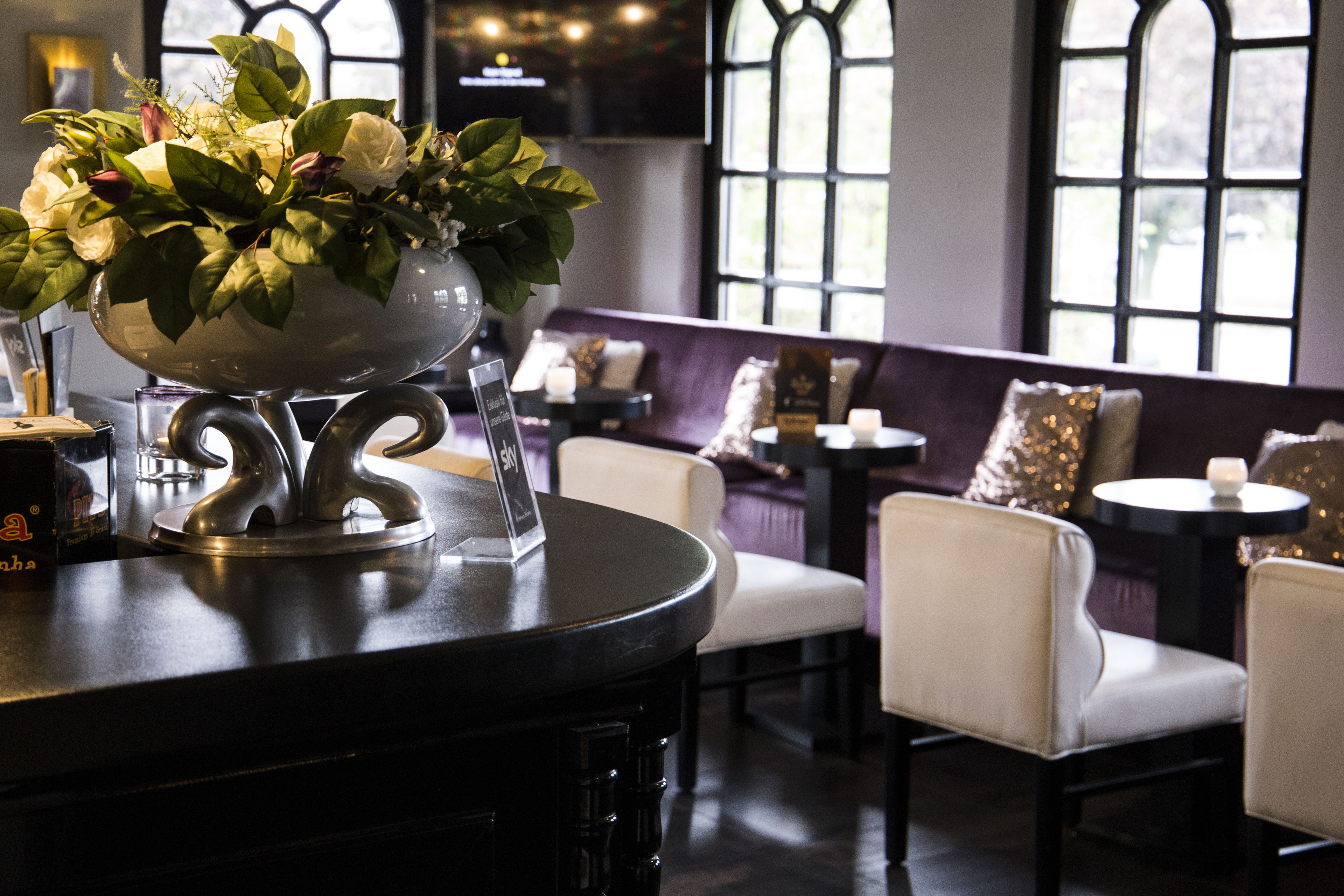 A bar area with a large silver bowl of flowers, a glass of water, and a sign reading "The Bar". There are white cushioned chairs and a small round table with a candle.