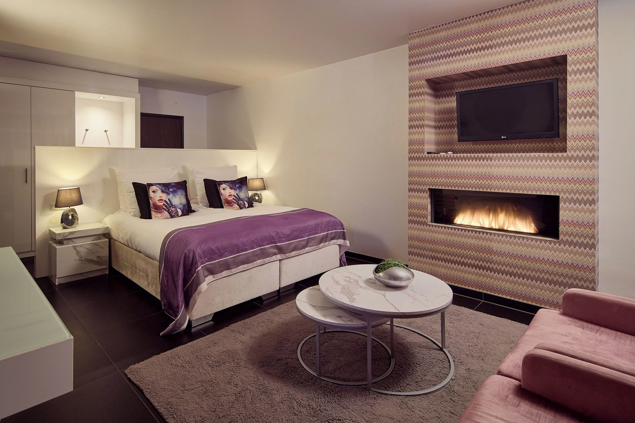 Een moderne slaapkamer met een bed, nachtkastjes, een ronde tafel, een roze bank, een open haard en een aan de muur bevestigde televisie.