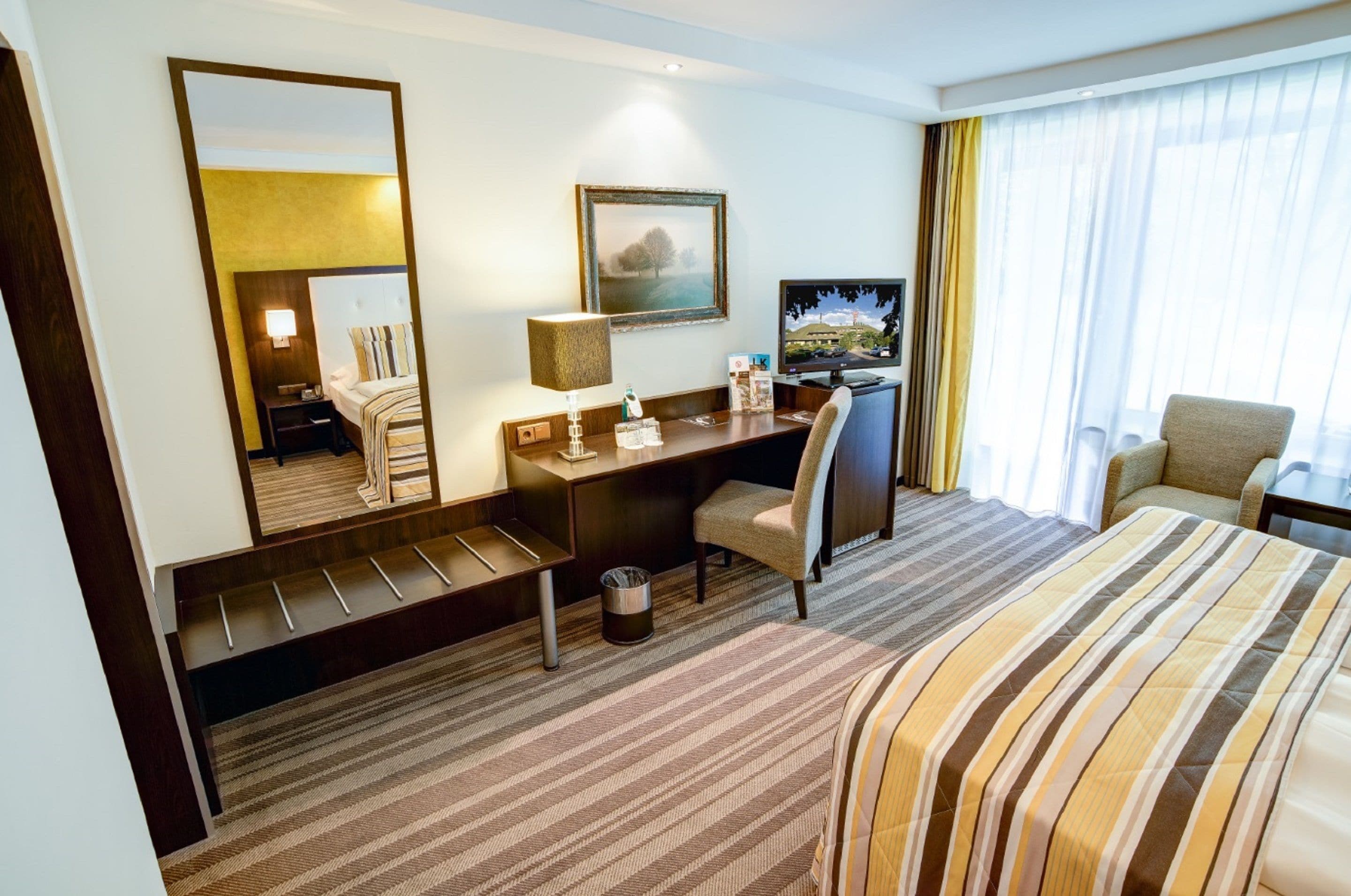 A hotel room with a bed, a desk, a chair, a television, a mirror, and a window with curtains.