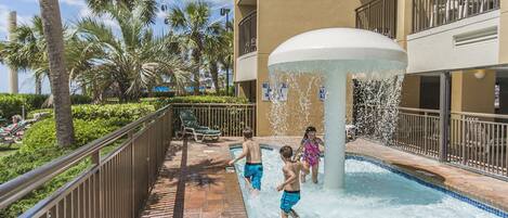 A pool area with a water fountain, palm trees, and a building with balconies.