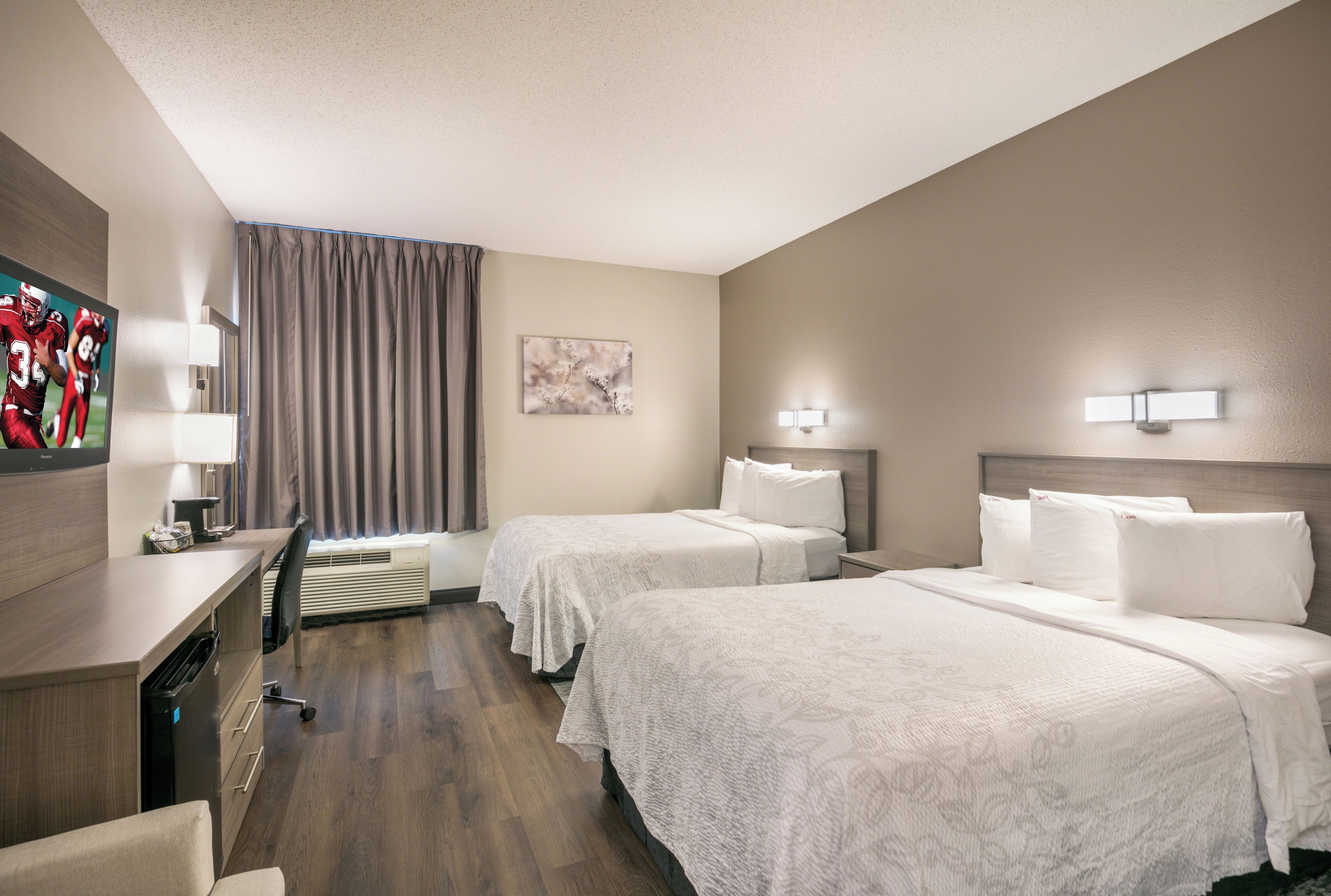 A hotel room with two beds, a desk, a chair, a flat-screen TV, and a window with curtains.