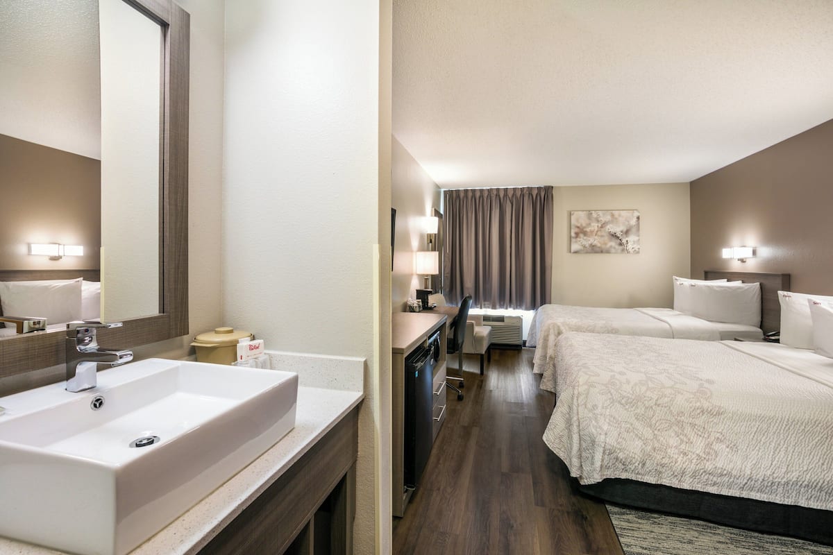 A hotel room with a large bathroom sink, a bed with a white comforter, a bedside table with a lamp, and a large mirror.