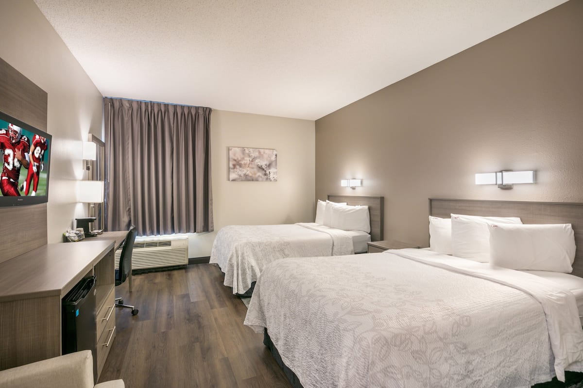 A hotel room with two beds, a desk, a chair, a flat-screen TV, and a painting on the wall.