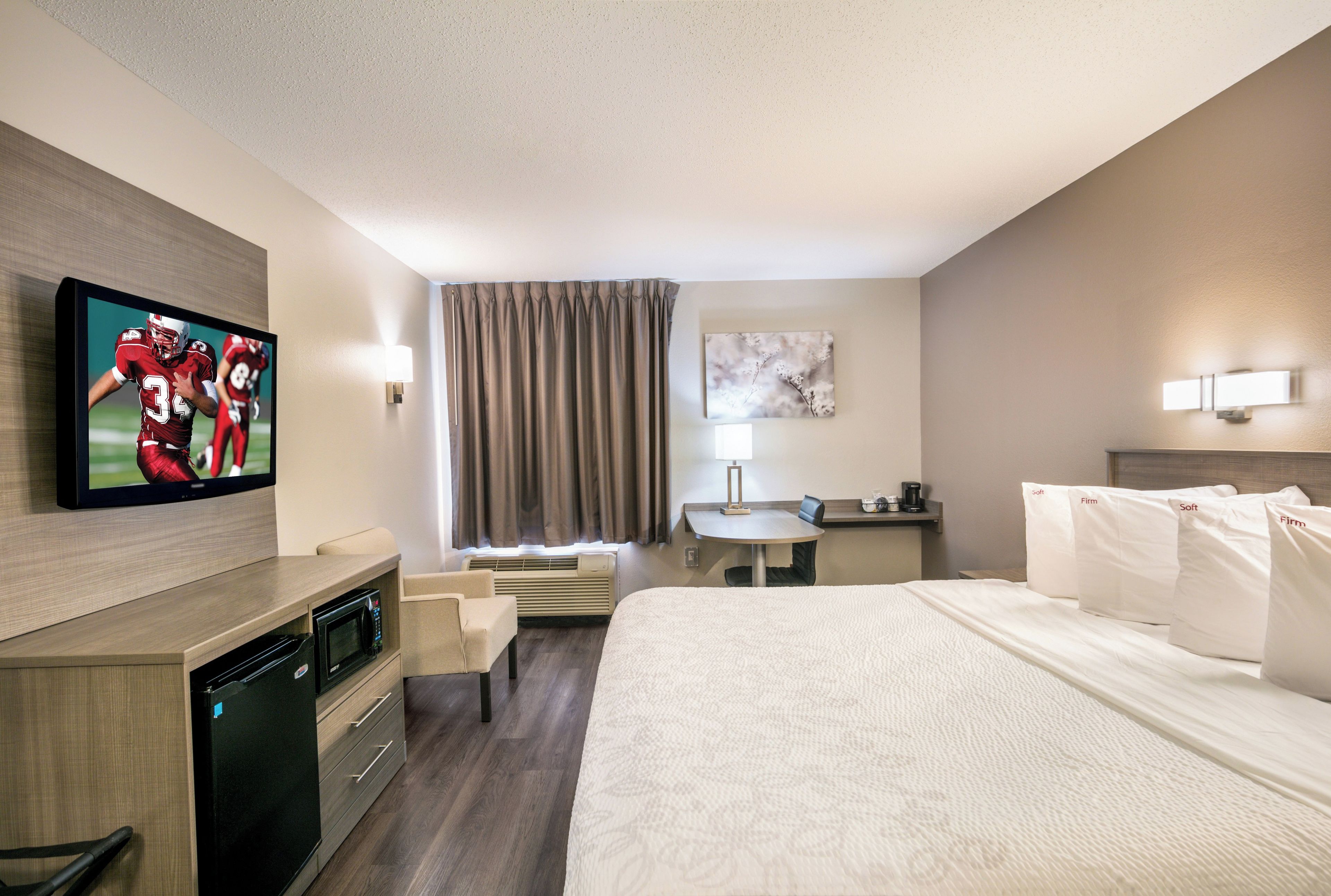 A hotel room with a flat-screen TV, a desk with a lamp, a bed with white bedding, and a wall-mounted artwork.