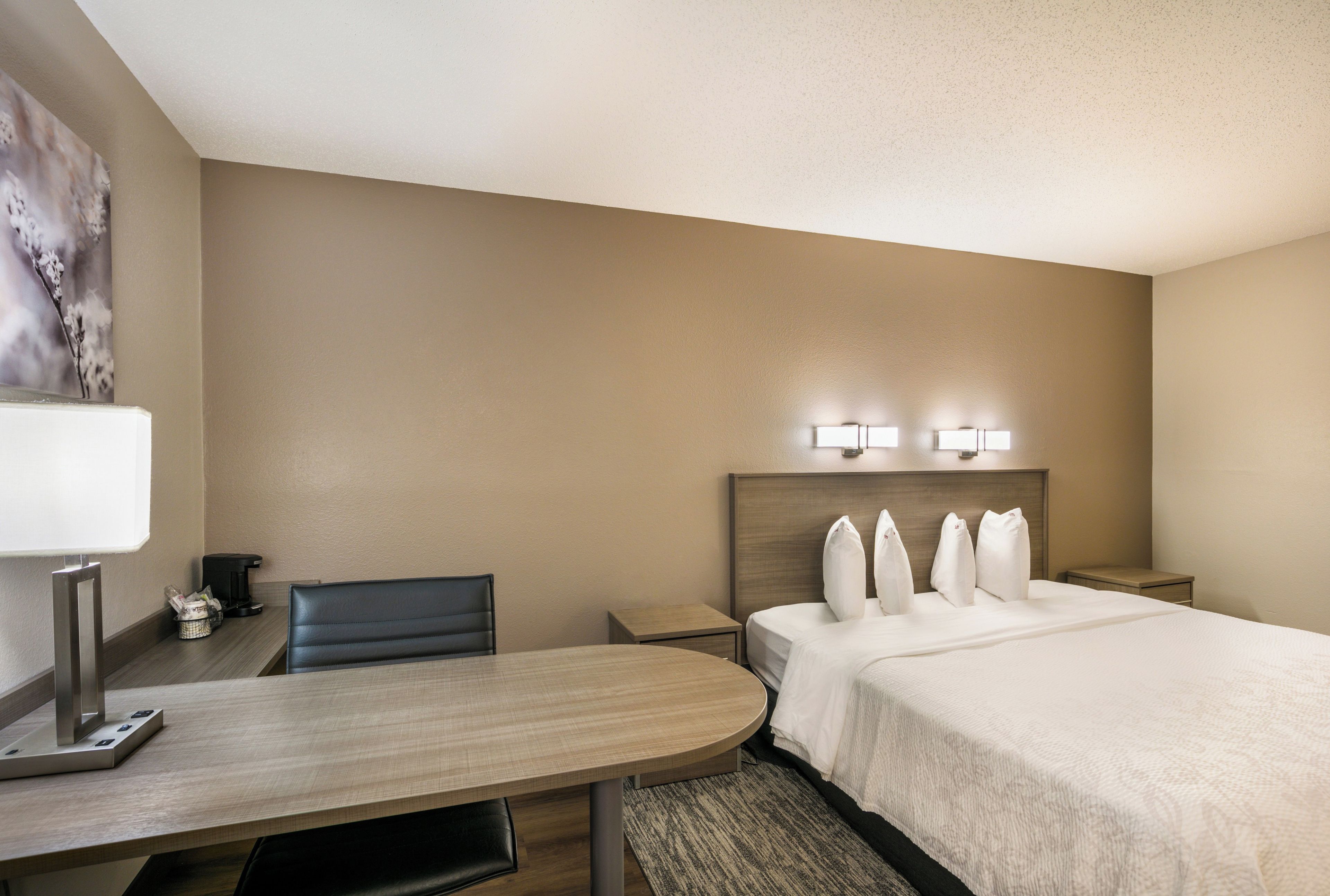 A hotel room with a bed, a desk, a chair, a nightstand, and a wall-mounted lamp.