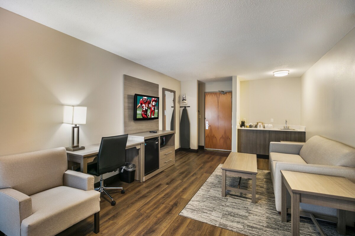 A hotel room with a flat-screen TV mounted on the wall, a desk with a chair, a sofa, a small table, and a kitchenette area.