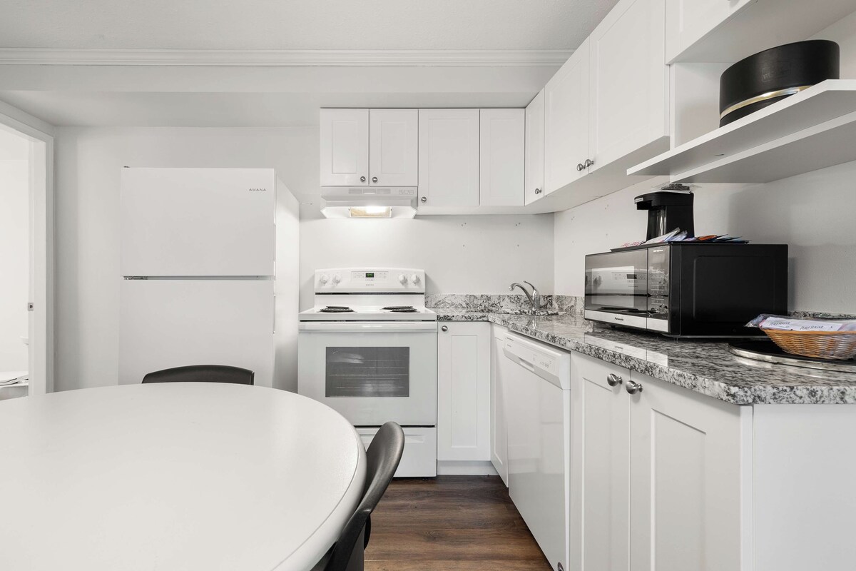 Une cuisine de taille réduite, avec des meubles blancs, un réfrigérateur blanc, une cuisinière, un four à micro-ondes et une machine à café.