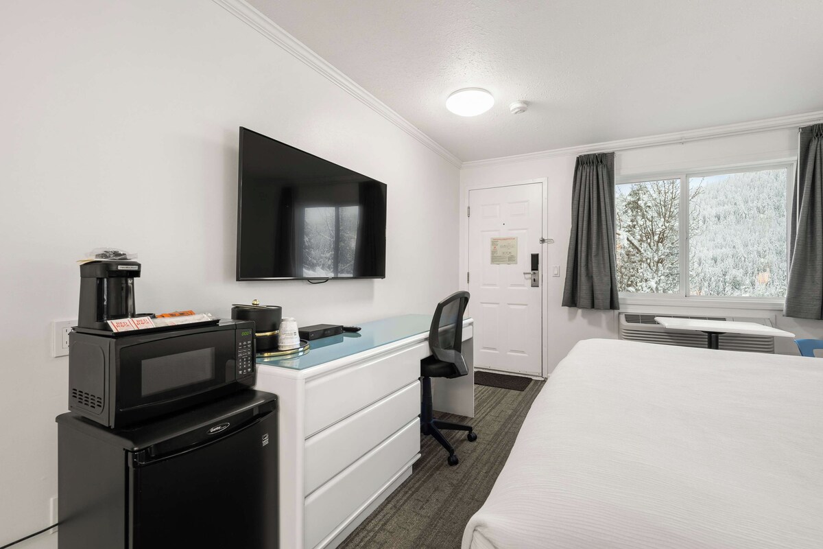 Une chambre d’hôtel équipée d’un lit, d’un bureau avec un ordinateur, d’un four à micro-ondes, d’une machine à café, d’une télévision et d’une fenêtre avec des rideaux.