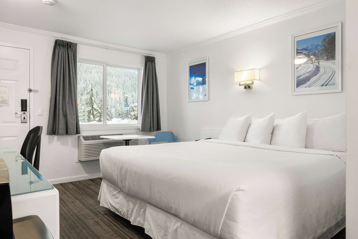 Une chambre d’hôtel avec un grand lit, un bureau avec une chaise, une fenêtre avec des rideaux, une lampe fixée au mur et un tableau encadré.