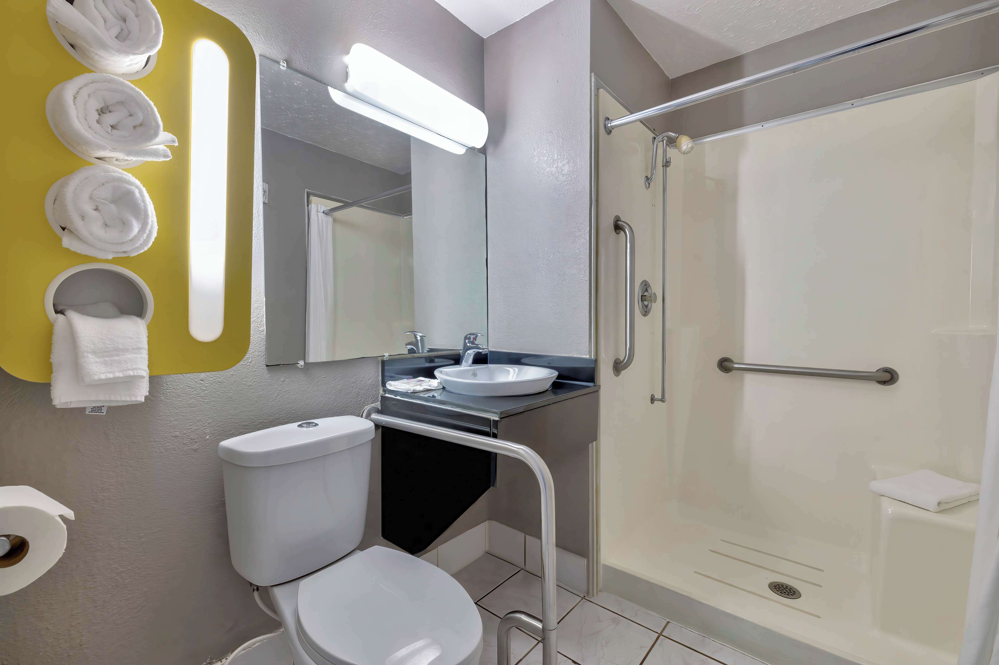 Ein Badezimmer mit Toilette, Waschbecken und Dusche. Handtücher hängen an der Wand und in der Dusche befindet sich ein Haltegriff.