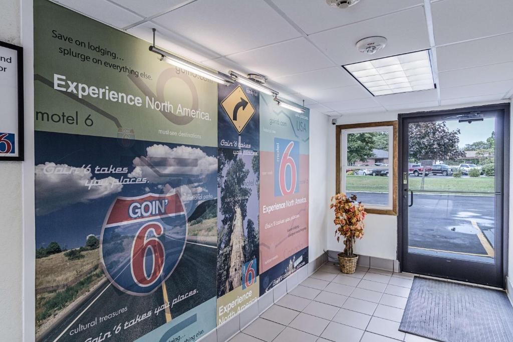 A hotel lobby with a large poster advertising the Goin' 6 experience in North America, a reception desk, and a view of the parking lot through the door.