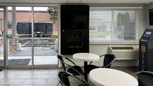 A modern indoor seating area with tables and chairs, a vending machine, and a view of the outside through a glass door. - Motel 6 Oshkosh, WI (Oshkosh)